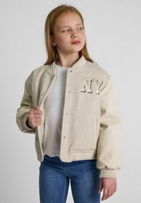 Bež jakna v varsity stilu z mehko teksturo, s pritisnimi gumbi in vezenino "NY". S trdo pletenicami na zapestjih in robu, nosi se over belo srajco.