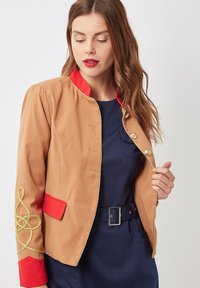 Mujer vestida con una chaqueta color beige con acentos rojos y bordados dorados sobre un vestido azul marino con un cinturón de hebilla cuadrada, mirando hacia abajo mientras sostiene la chaqueta abierta.