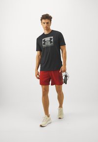 Svart kortermet t-skjorte med grå Under Armour-logo, kombinert med røde treningsshorts og beige sneakers. Hoppetau i hånden.