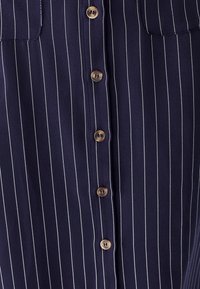 Tissu bleu marine à fines rayures verticales blanches, avec une rangée centrale de six boutons marron à quatre trous chacun.