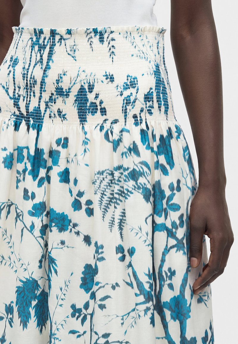 Ansicht von der Taille bis zur Mitte des Oberschenkels einer Person, die einen weißen Rock mit blauen Blumen- und Farnmustern und einem gerafften Bund trägt, kombiniert mit einem weißen Oberteil.