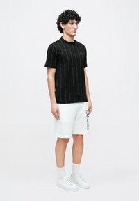 Férfi áll, fekete rövid ujjú pólót visel, amelyen függőleges felirat van, fehér rövidnadrágot fekete oldalsó feliratokkal, fehér zoknit és fehér tornacipőt.