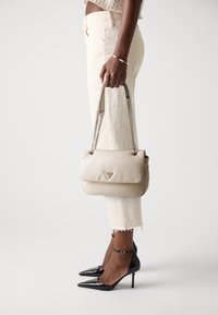 Borsa a tracolla in pelle beige con chiusura a patta, hardware dorato e tracolla a catena. Indossata con pantaloni bianchi a tre quarti e décolleté neri a punta.