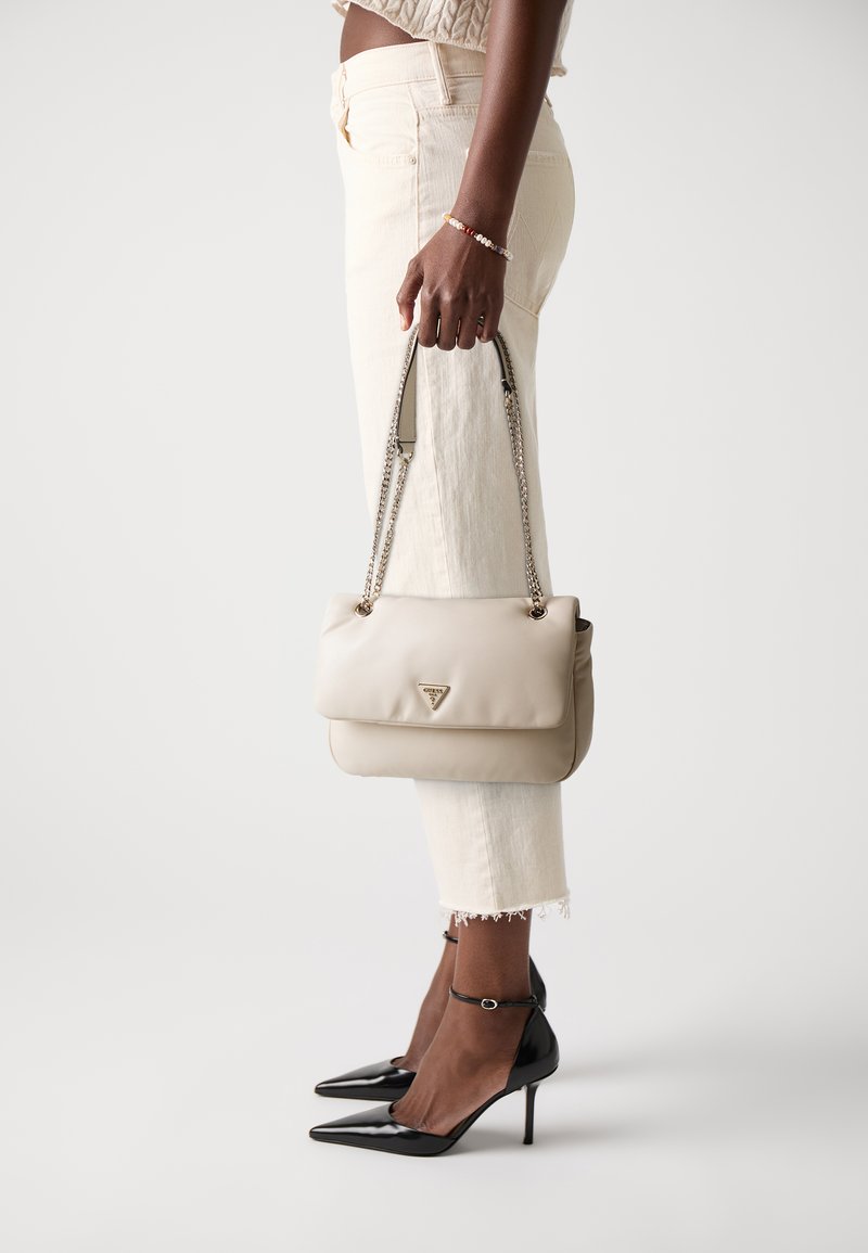 Borsa a tracolla in pelle beige con chiusura a patta, hardware dorato e tracolla a catena. Indossata con pantaloni bianchi a tre quarti e décolleté neri a punta.