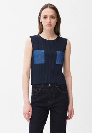 Femme portant un haut noir sans manches en maille côtelée avec deux poches carrées en denim bleu sur la poitrine et un jean noir taille haute, sur fond blanc.
