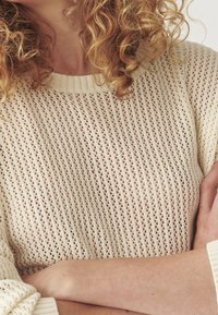 Strickpullover in Creme, mit einem lockeren, offenen Maschenmuster und einem gerippten Ausschnitt. Lange Ärmel und geraffte Akzente an den Säumen sind gut sichtbar.