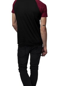 Černé tričko s krátkým rukávem a maroon raglánovými rukávy, v kombinaci s černými zúženými džínami. Výrazné tetování na pravém předloktí.