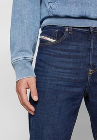 Dark blue denim jeans with a slight sheen, contrasting yellow stitching, and a subtle textured finish. Hand resting on the thigh area.