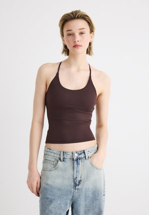 Tânără purtând un top maro fără mâneci și blugi albaștri deschiși, stând în picioare pe un fundal alb simplu.