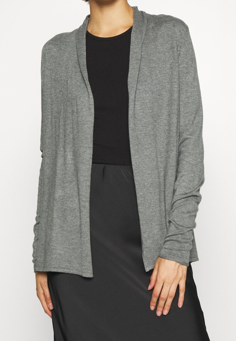 Personne portant un cardigan gris à manches longues et ouvert sur le devant, au-dessus d'un haut noir et d'une jupe noire, debout avec une main détendue le long du corps.