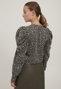 Sort og beige puffærmet bluse med blomstermønster, struktureret stof, rund halsudskæring og manchetter med knapper, sammen med en mørk nederdel.