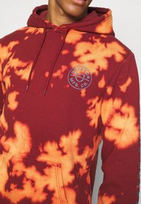 Felpa con cappuccio tie-dye bordeaux e arancione realizzata in cotone, con tasca frontale e logo blu sul petto. Cappuccio con coulisse e texture morbida.
