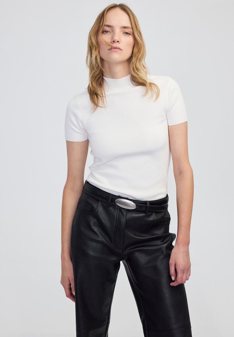 Blonde Frau trägt ein weißes kurzärmliges Top mit Stehkragen, das in eine hochtaillierte schwarze Lederhose mit einem schwarzen Gürtel und ovaler Schnalle gesteckt ist.