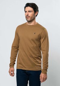 Homme aux cheveux foncés et à la barbe naissante, portant une chemise marron à manches longues et un jean bleu foncé, debout devant un fond clair uni.