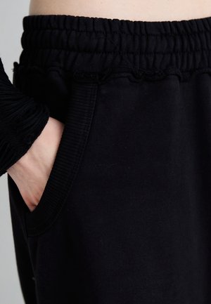 Persona con pantalones negros de cintura elástica, mano izquierda en el bolsillo delantero, torso parcialmente visible sobre un fondo neutro.