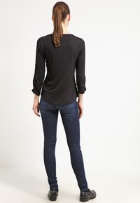 Žena s hnědým ohonem, oblečená v černém tričku s dlouhým rukávem, tmavomodrých skinny džínech a černých botách, stojící čelem k bílému pozadí.