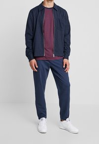 Chaqueta de rayas azul marino con cremallera, camiseta de rayas rojas debajo y pantalones azul marino. Zapatillas blancas completan el conjunto. Diseño casual y relajado.