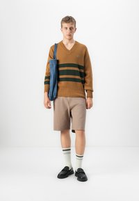 Young man wearing brown V-neck sweater with green stripes, beige shorts, white striped socks, black shoes, and carrying blue shoulder bag.