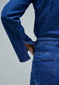 Veste en denim bleu foncé avec des manches longues et un ourlet arrondi surpiqué. Le dos présente une passante de ceinture et un détail de poche arrière.