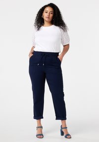 T-shirt blanc avec des manches perforées, associé à un pantalon bleu marine tapered avec des poches latérales et une taille à cordon. Sandales à talons bleues.