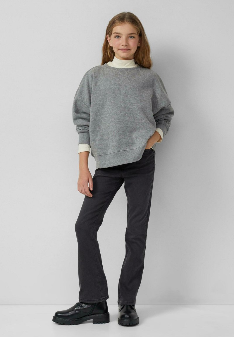 Felpa oversize grigia chiara con spalle scese, indossata sopra un dolcevita bianco, abbinata a jeans scuri slim-fit e stivali lacci neri.