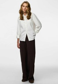 Bijela košulja od pamuka s dugim rukavima i opuštenim krojem u kombinaciji s širokim tamnobraon hlačama.