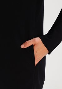 Černý oděv s rukou v postranní kapse. Materiál se zdá být hladký, s dlouhými rukávy a volným střihem, který zdůrazňuje jednoduchost.