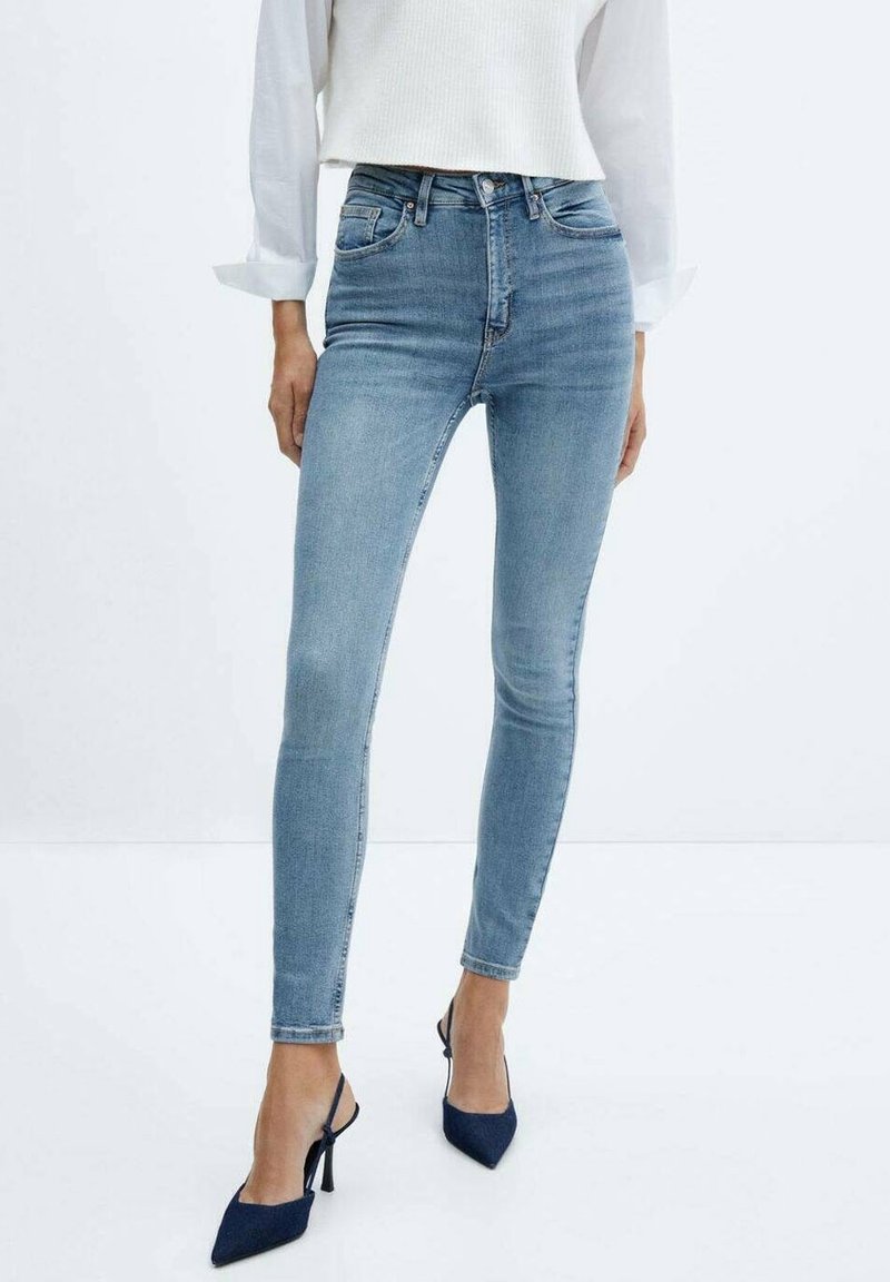 Femme portant un jean skinny bleu clair, une chemise blanche à manches longues et des escarpins slingback pointus bleu marine, debout sur un sol blanc.