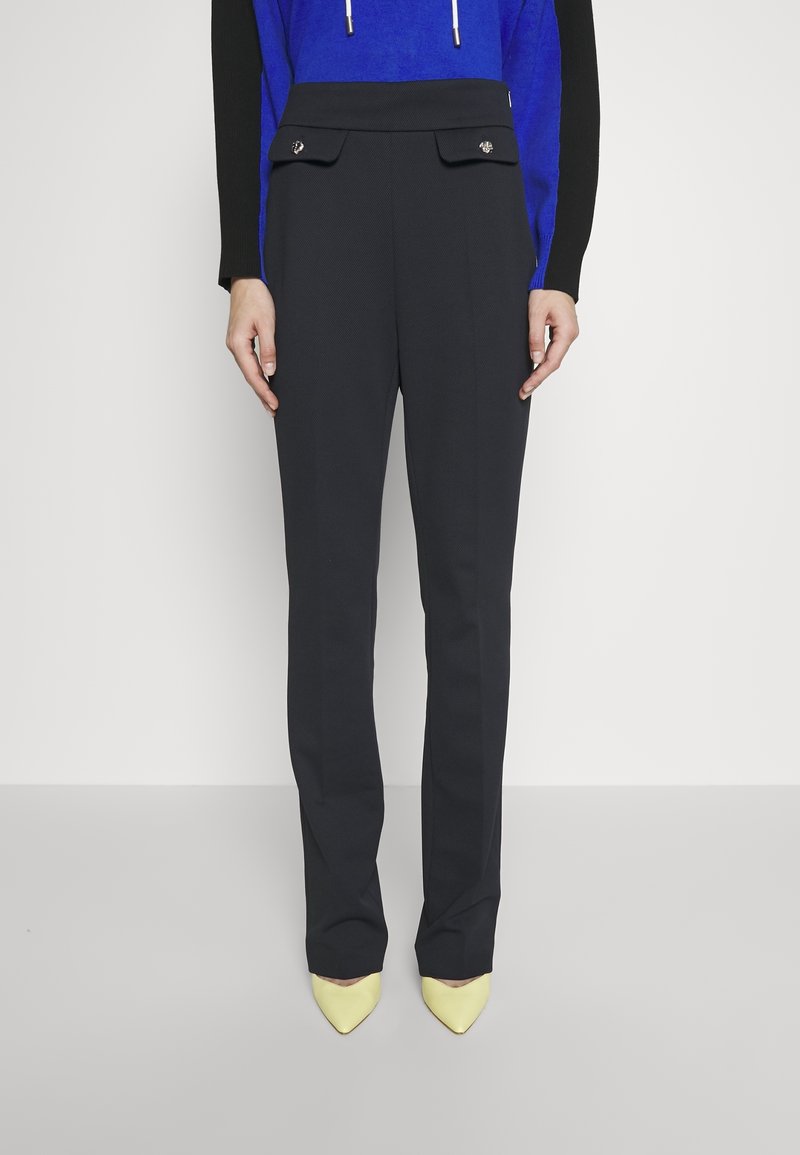 Pantalons ajustés bleu marine à taille haute, dotés de poches avant doubles et de boutons décoratifs, associés à des talons jaunes pointus.