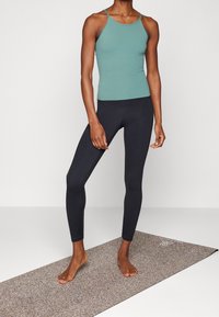 Top de entrenamiento en color teal claro combinado con leggings negros, fabricado con un tejido elástico. Esteras de yoga texturizada en color marrón debajo.