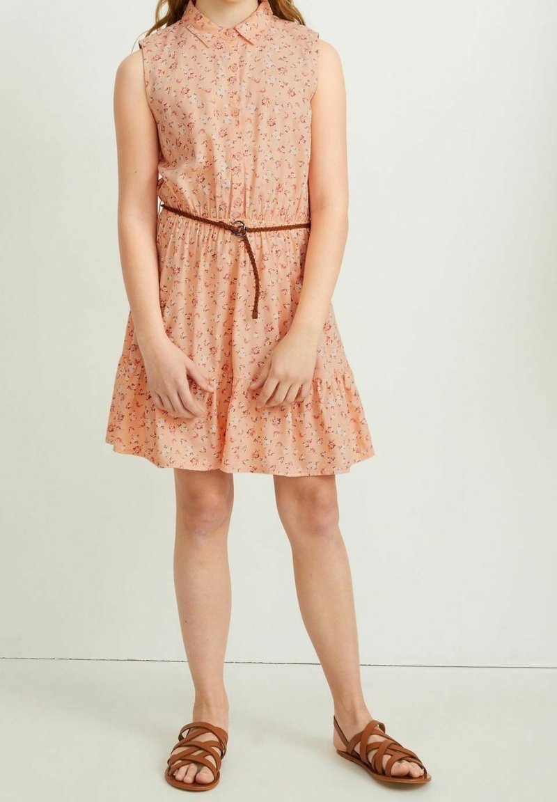 Fille portant une robe sans manches pêche à motifs floraux, avec une fine ceinture marron et des sandales marron à brides, debout sur un fond uni.