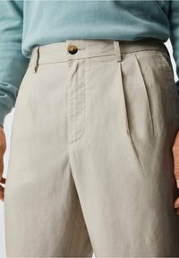 Beige plissébroek met een ritssluiting aan de voorkant en een bruine knoopsluiting, gemaakt van lichte stof met een gladde textuur.