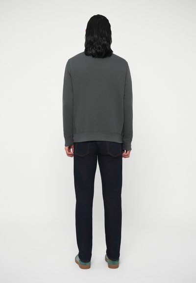 Vyras stovi atsukęs nugarą, apsirengęs tamsiai pilku džemperiu, tamsiai mėlynomis džinsomis ir melsvais sportbačiais priešais paprastą baltą foną.