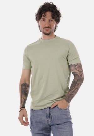 Uomo con capelli ricci scuri, maglietta verde chiaro, jeans blu e tatuaggi su tutto il braccio, in piedi con una mano in tasca davanti a uno sfondo uniforme.