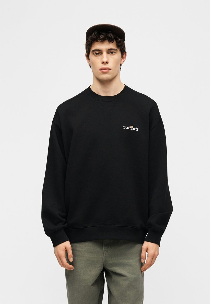 Giovane uomo che indossa una felpa nera Carhartt e pantaloni verde oliva, in piedi davanti a uno sfondo chiaro e uniforme.