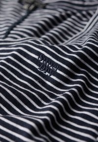 Mørkeblå og hvide stribede stof med broderet "Super Dry"-logo i matchende mørkeblå tråd, vist tæt på med synlig tekstur og folder.