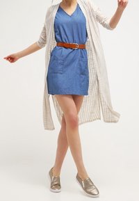 Džínové šaty s krátkým rukávem a předními kapsami, doplněné béžovým pruhovaným kardigánem a metalickými nazouvacími botami. Hnědý pásek zvýrazňuje pas.