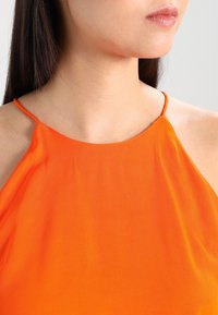 Top arancione senza maniche con spalline sottili, realizzato in tessuto a costine con una texture liscia. Il colletto è rotondo, mostrando una vista ravvicinata.