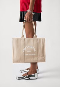 Borsa tote in tela beige con due robuste maniglie, con testo bianco: "KARL LAGERFELD PARIS 21 RUE ST-GUILLAUME." Scarpe basse lisce argentate.