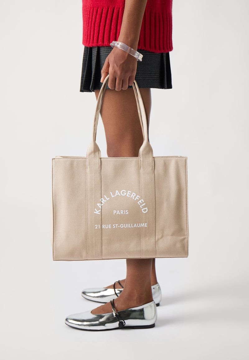 Borsa tote in tela beige con due robuste maniglie, con testo bianco: "KARL LAGERFELD PARIS 21 RUE ST-GUILLAUME." Scarpe basse lisce argentate.