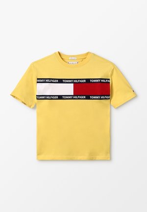 T-shirt in cotone giallo con maniche corte, con una banda orizzontale con la scritta "TOMMY HILFIGER" in nero, bianco e rosso.