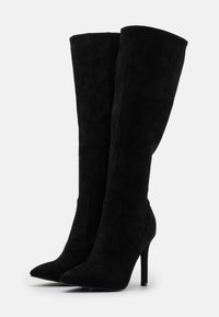 Botas negras por encima de la rodilla hechas de ante, con punta afilada, diseño curvado y tacón de aguja. Textura lisa con detalles mínimos.