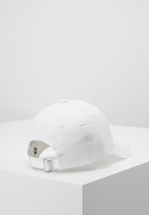 White adjustable baseball cap with metal buckle strap, resting on a white corner surface against a plain light gray background.