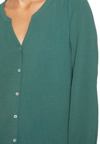Camicia verde abbottonata con scollatura a V, maniche lunghe e tessuto testurizzato. Presenta piccoli bottoni rotondi e una vestibilità comoda.