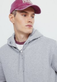 Szary hoodie z zamkiem z przodu, sznurkami i miękką teksturą, nałożony na biały i bordowy t-shirt. Nosi jaskrawofioletową czapkę.