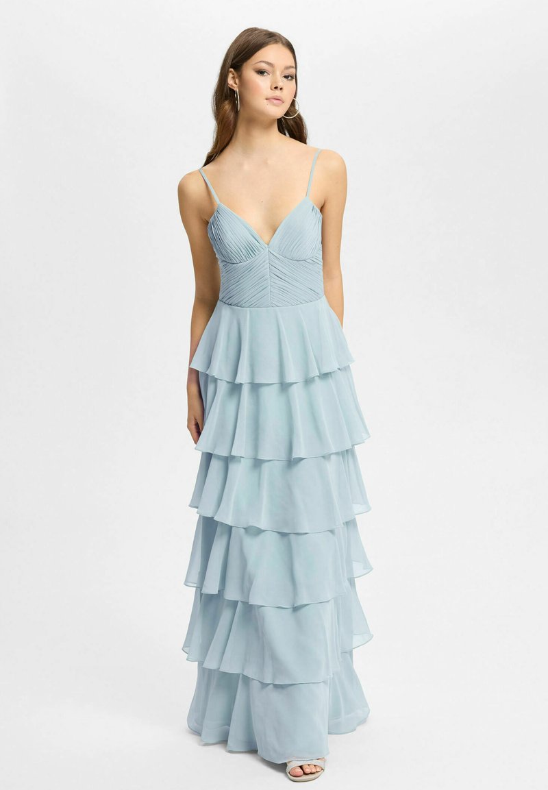 Femme portant une robe bleu clair sans manches avec une jupe à volants superposés et des bretelles fines, posant devant un fond blanc uni.