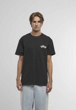 Mladý muž so svetlohnedými vlasmi, oblečený v čiernom tričku s nápisom "UNFAIR" a svetlomodrých džínsoch, stojaci pred jednoliatym sivým pozadím.