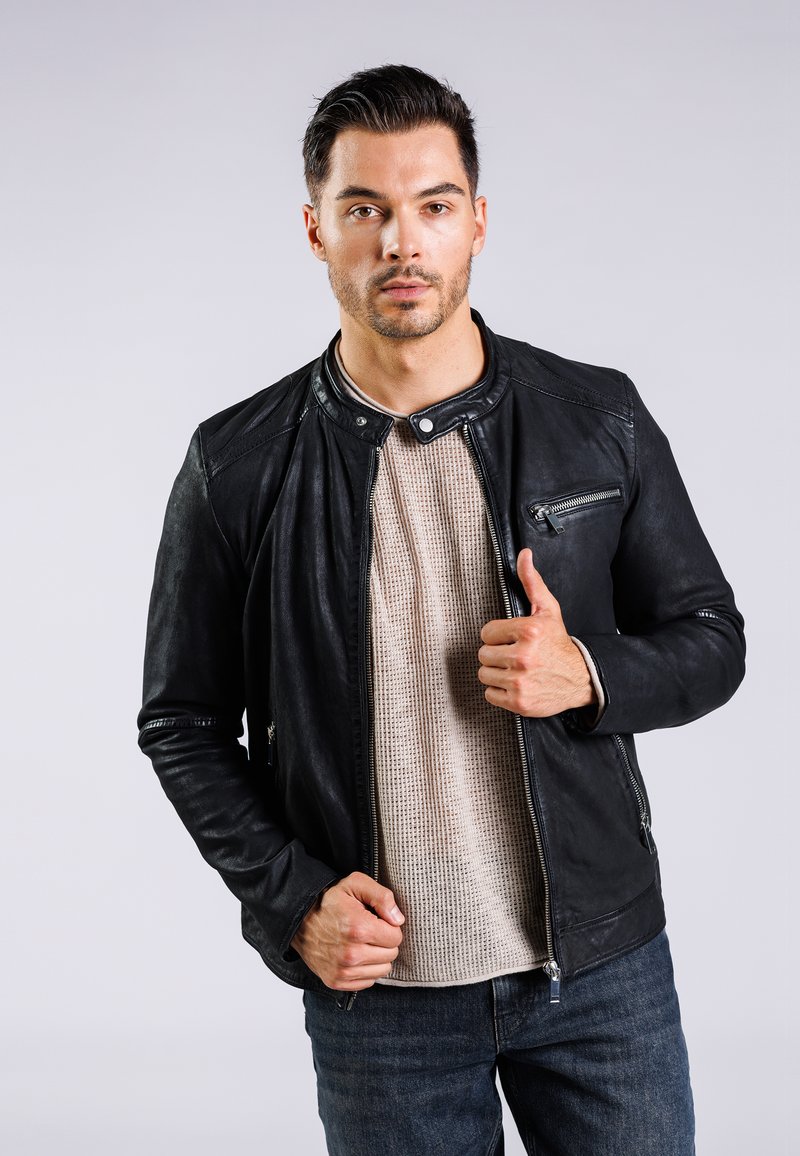 Chaqueta de motocicleta de cuero negro con un bolsillo en el pecho con cremallera, collar a presión y detalles texturizados, superpuesta sobre una camiseta de punto beige.