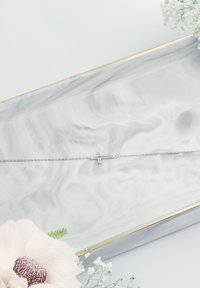 Bracciale in argento con un semplice pendente a croce. Superficie liscia con finitura lucida, esposto su un vassoio marmorizzato con accenti floreali.