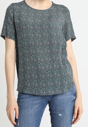 Kobieta nosi ciemnozieloną bluzkę z krótkim rękawem w małe białe i turkusowe kwiatowe wzory, połączoną z niebieskimi podniszczonymi dżinsami.